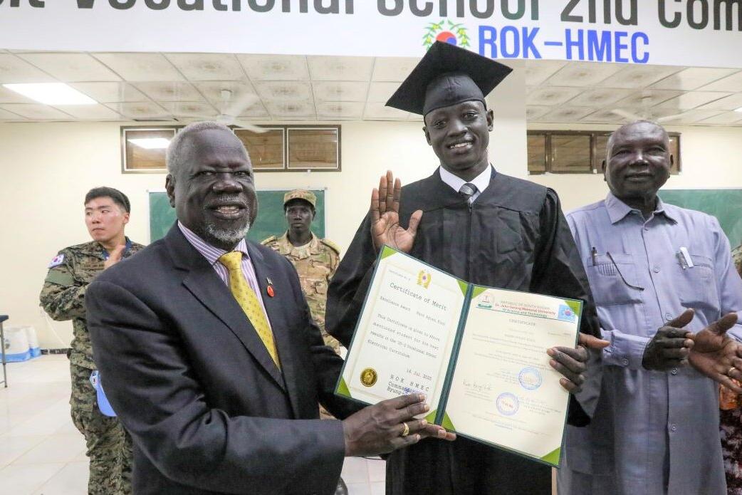 Graduation ceremony indoors with a person in a cap and gown holding a certificate, surrounded by others, with a banner for a vocational school visible above.