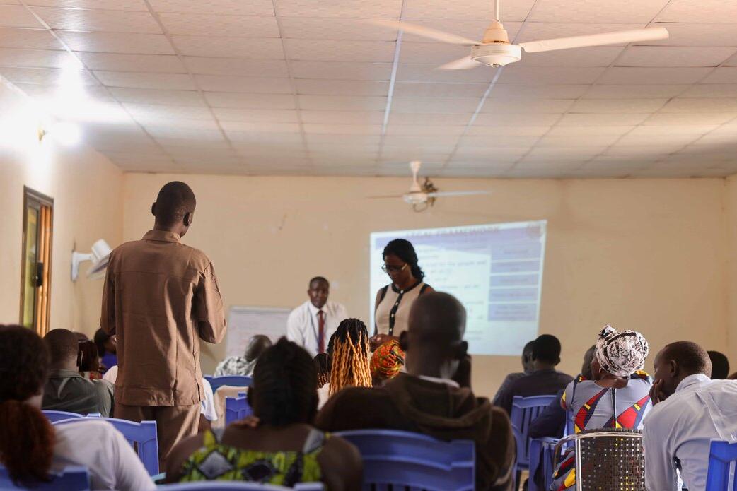 A classroom setting with people seated on blue chairs facing a projector screen, while two presenters stand at the front and one participant stands to speak.