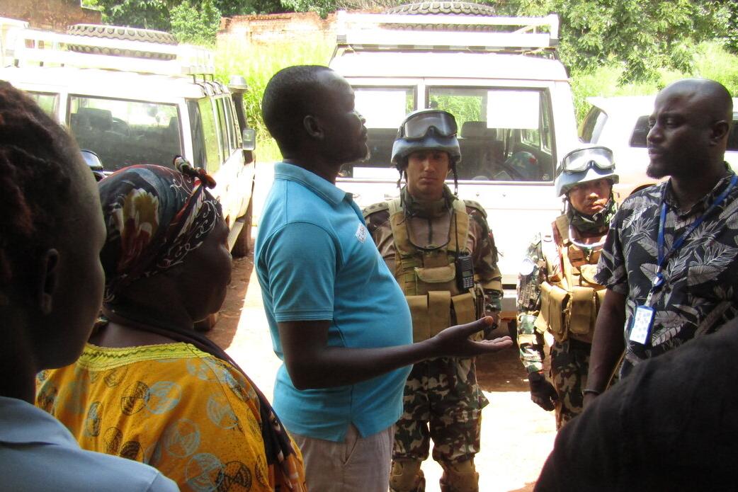 Small group of people standing and talking near parked vehicles, including two individuals in camouflage uniforms and protective gear.
