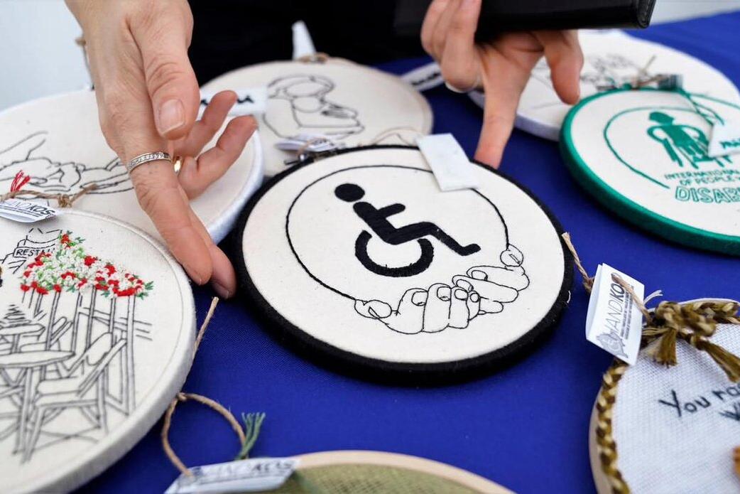 Close-up of embroidered circular patches displayed on a blue surface. One patch in the center shows a wheelchair accessibility symbol held by two hands. Other patches feature various designs, including flowers and text.