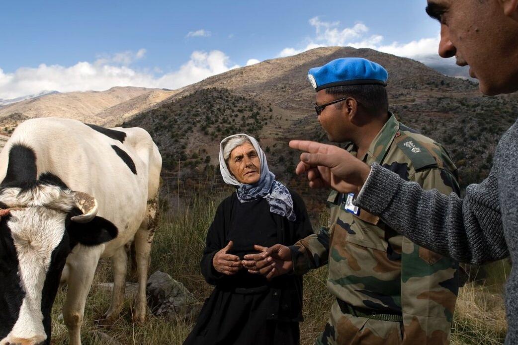 A peacekeeper speaks with two individuals on rural land beside a cow.