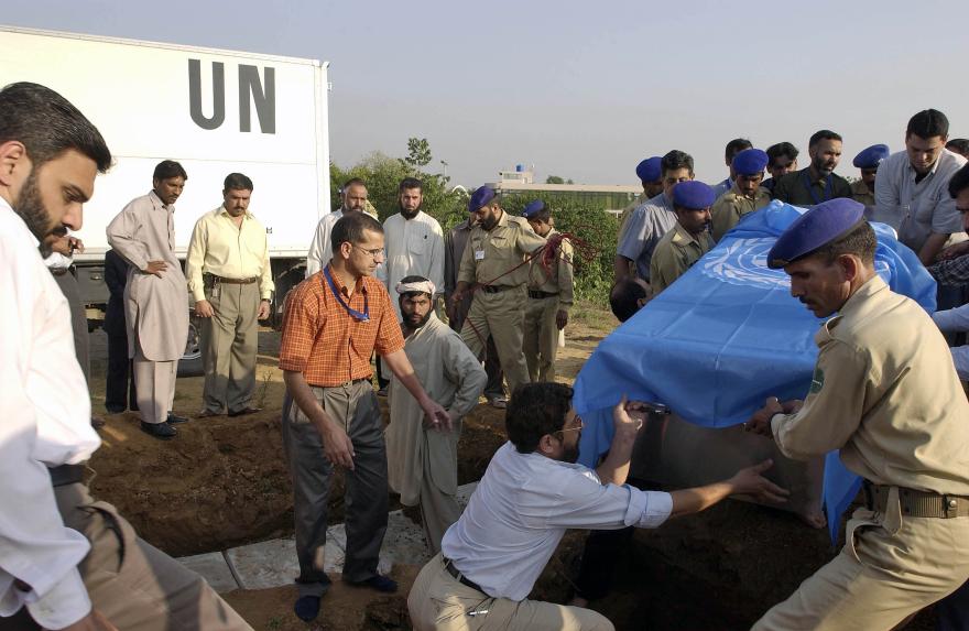 Group of people lowering a coffin draped in a UN flag into a grave, with a UN truck in the background.